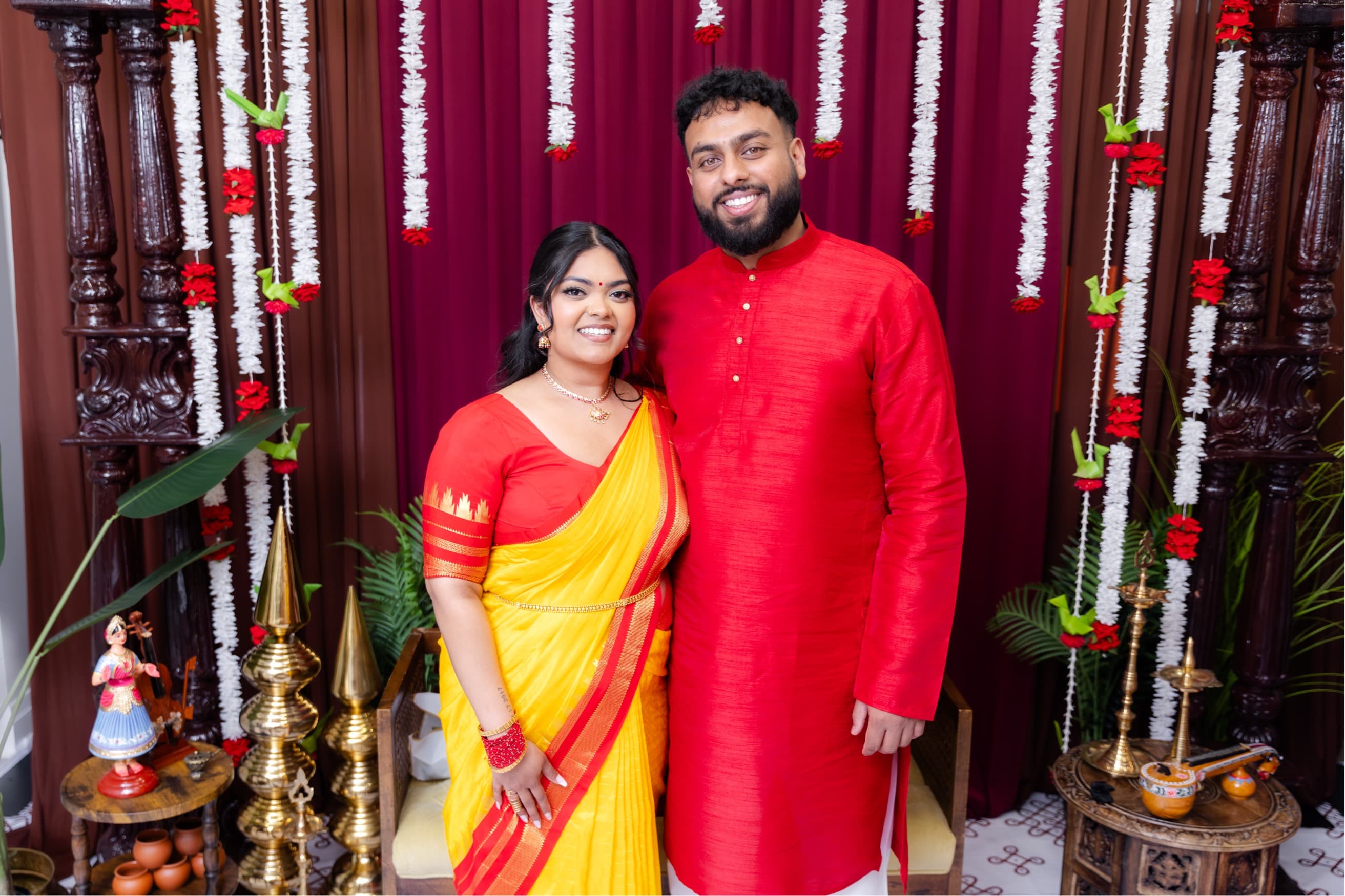 Piratheepan and Shivanna smiling together in front of a maroon curtain decorated with white and red floral garlands, Shivanna in a yellow and red saree and Piratheepan in a red kurta
