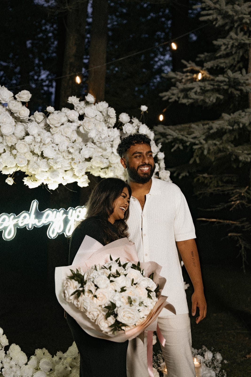 Piratheepan and Shivanna embracing in front of a white rose floral arch lit with a neon sign and string lights during the proposal, Shivanna holding a large white bouquet